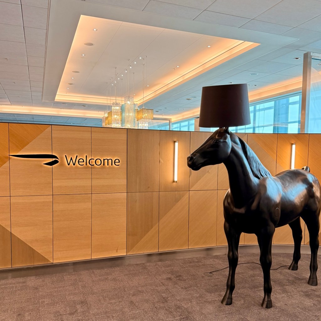 An interior view of British Airways' Galleries First Lounge featuring a black horse statue with a lamp as its head, next to a wood-paneled wall with a 'Welcome' sign.