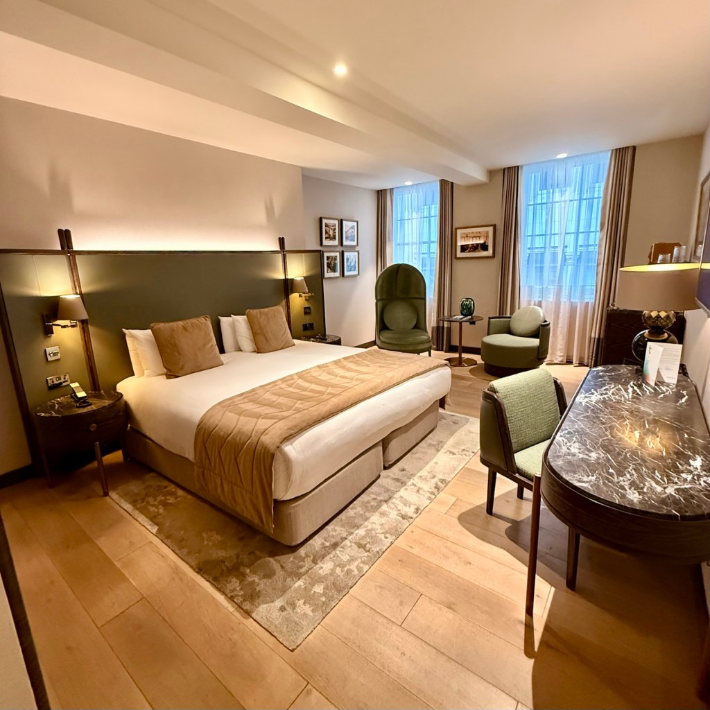 A luxurious hotel room featuring a spacious bed with plush bedding, elegant armchairs, a stylish writing desk, and large windows allowing natural light to fill the space.