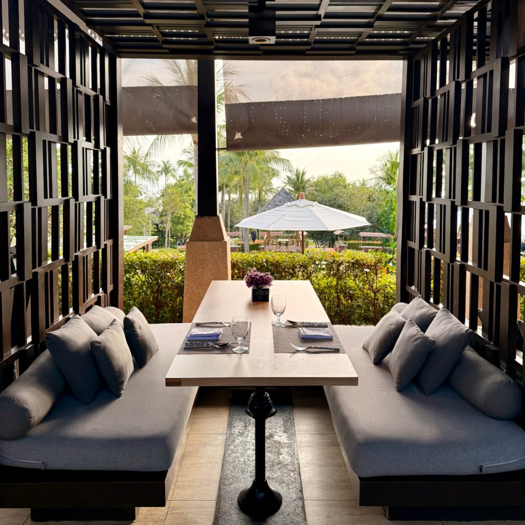 A stylish dining area with comfortable seating, showcasing a table set with glassware and tableware, and a view of palm trees and an outdoor umbrella in the background.