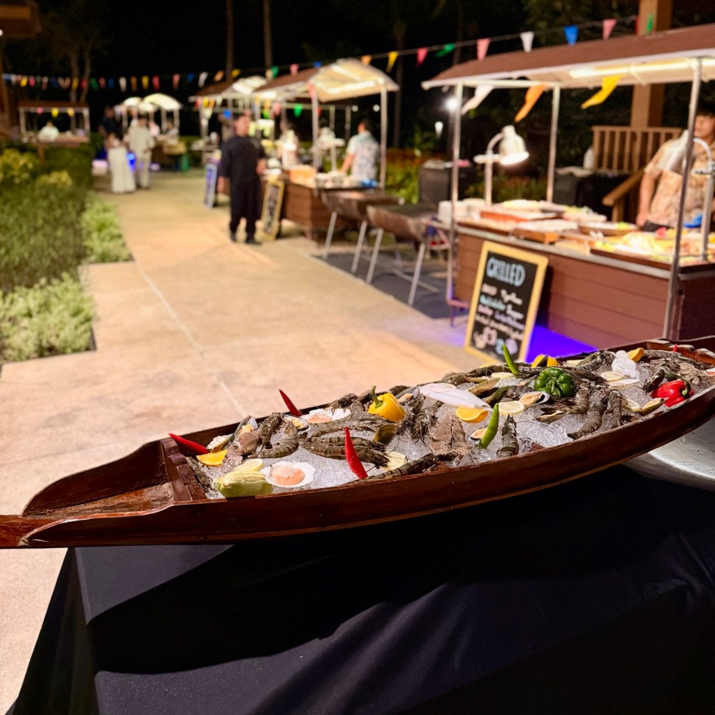 A wooden boat filled with seafood, including lobsters and garnished with lemon slices and chilies, displayed at an outdoor dining event with food stalls in the background.